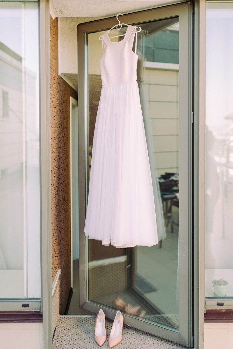 Chic white wedding gown elegantly hung outside a glass door with matching shoes.