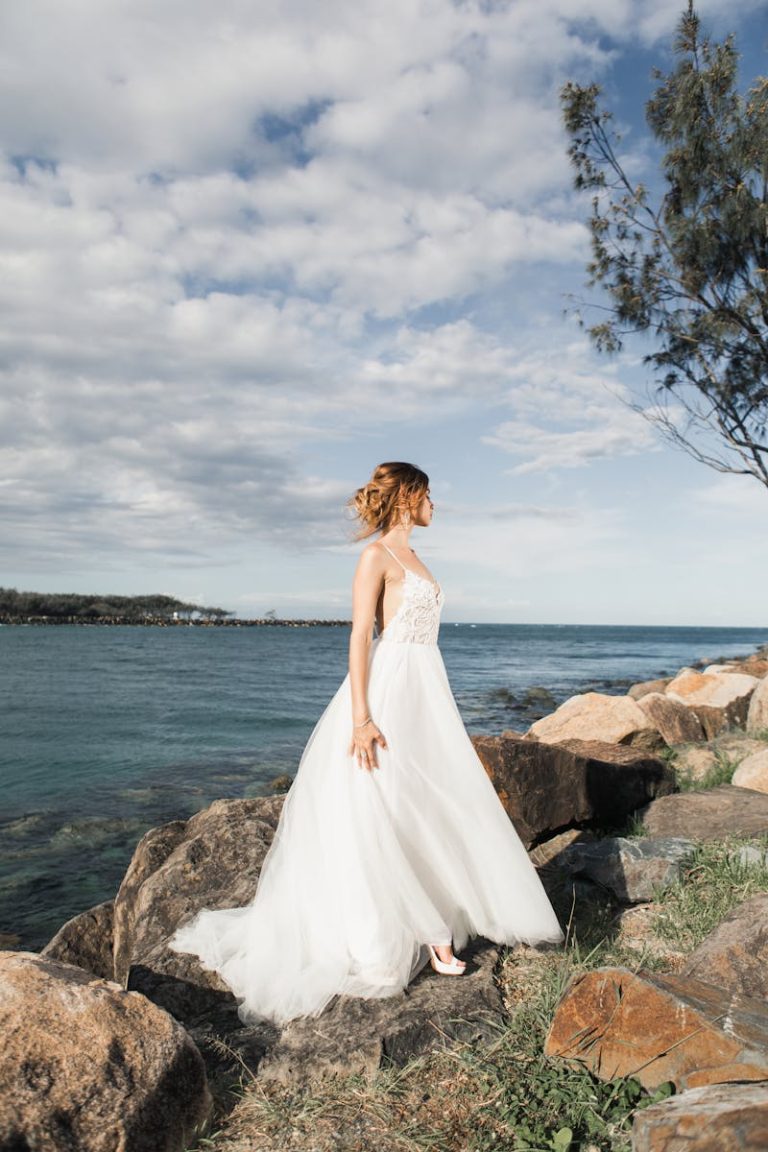 A bride in an elegant white gown stands on rocks by the ocean, gazing at the horizon under a clear sky.