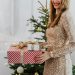 Festive scene with smiling woman in a sparkly dress holding wrapped presents by a decorated Christmas tree.