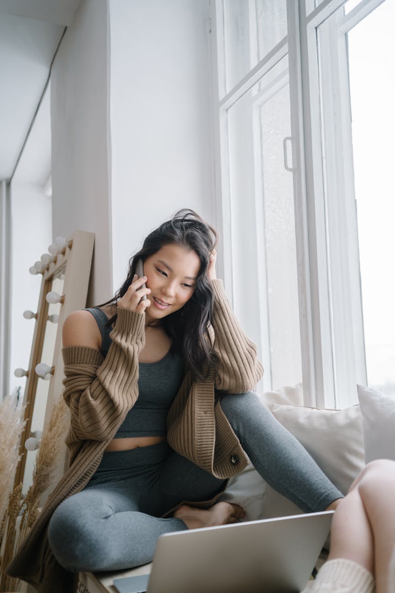 A woman sits comfortably at home, chatting on her phone near a large window.