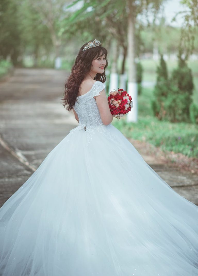 Beautiful bride in an elegant white wedding gown holding a red bouquet outdoors.