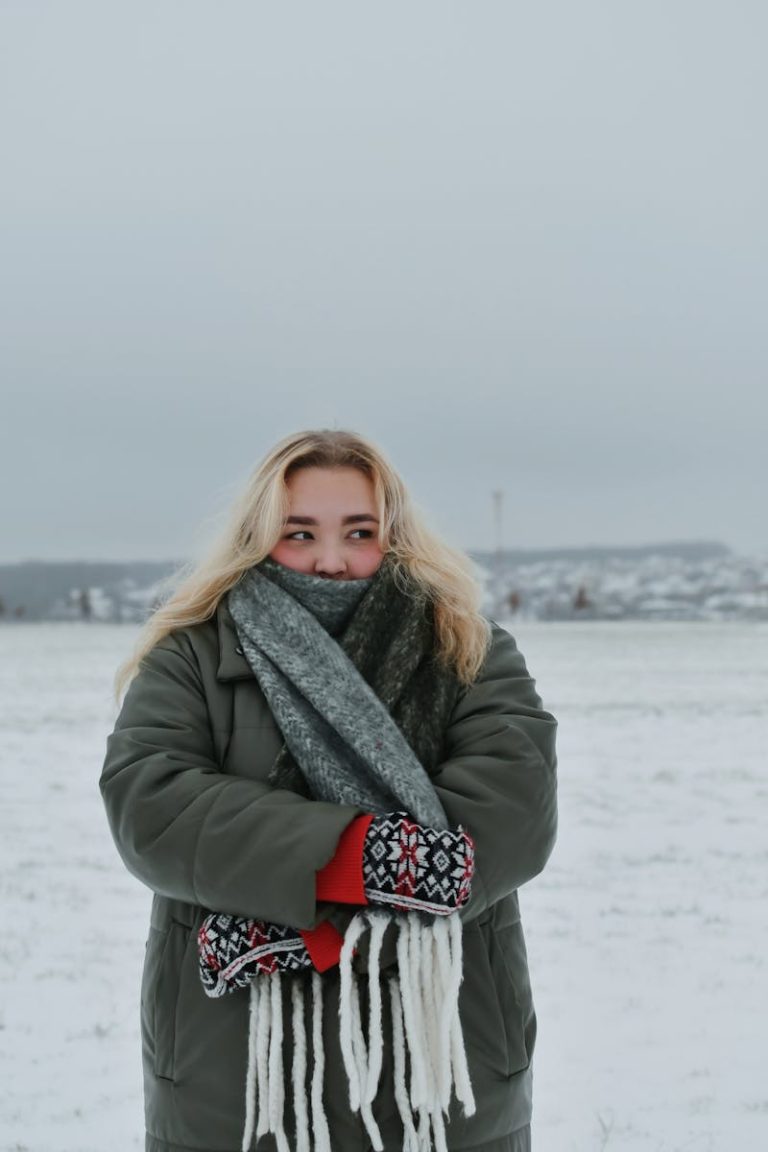 A woman bundled in winter clothing with a scarf and gloves stands outdoors in a snowy field.