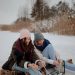Two women smiling while tying ice skates on a frozen lake in winter attire.