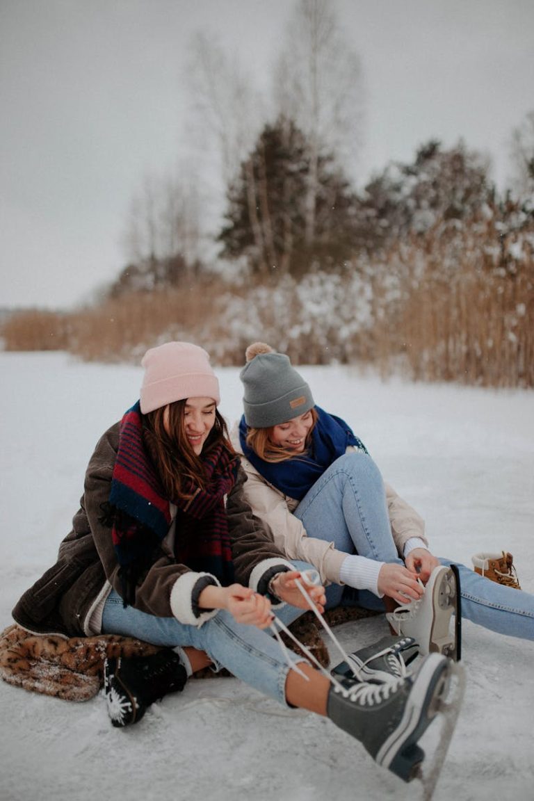 Two women smiling while tying ice skates on a frozen lake in winter attire.